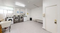 Located at 46 Saranac in Toronto's M6A neighbourhood, this shared laundry facility is a practical and well-maintained space designed to serve building residents efficiently. The room is finished with light grey ceramic tile flooring throughout, contributing to a clean and cohesive aesthetic that is easy to maintain.

The laundry area is equipped with multiple front-loading washing machines and dryers, providing ample capacity for regular use. A freestanding utility sink with a tiled backsplash offers added convenience for hand-washing or pre-treating garments. A white folding table provides a dedicated surface for sorting and folding laundry, enhancing the functionality of the space.

Bright fluorescent overhead lighting ensures the room is well-lit at all times. A window along one wall allows for natural light to supplement the artificial lighting, keeping the atmosphere open and pleasant. The walls are finished in crisp white, reinforcing the clean and orderly environment.

Practical amenities include a notice board displaying building information and instructions, a fire extinguisher for safety compliance, a waste bin, and a hand sanitizer dispenser near the entrance. A security camera is also present, adding an element of safety for users. The space is accessed through a solid entry door, ensuring a secure and contained environment for residents.