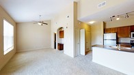This single-story residence showcases an open-concept layout that seamlessly connects the living area, dining nook, and kitchen. The living room immediately impresses with its vaulted ceilings that create a generous sense of vertical space, complemented by a ceiling fan with integrated lighting. Plush, neutral-toned wall-to-wall carpeting extends throughout the main living area, contributing to a warm and comfortable atmosphere.

A distinctive arched doorway serves as a decorative architectural detail, framing the transition between the living space and an adjoining area that features warm wood-toned cabinetry, ideal for use as a built-in desk or dry bar. Multiple doors visible from the main living area suggest access to additional rooms, closets, or utility spaces, adding functional versatility to the floor plan.

The kitchen is partially visible and features a raised countertop bar, dark laminate countertops, and a standard refrigerator, providing an efficient workspace that remains visually connected to the main living area. The neutral beige wall tones and multiple vents throughout reflect a practical, well-ventilated design, making this a comfortable and functional single-floor living space.