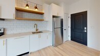 This single-floor unit showcases a contemporary design with light hardwood flooring throughout, crisp white walls, and recessed lighting that create a bright, clean aesthetic. The open-concept living area is generously proportioned, featuring a ceiling fan, a wall-mounted fireplace, and large black-framed windows that invite abundant natural light and offer views of the surrounding urban streetscape. A private balcony is accessible from the main living space, extending the area outdoors.

The kitchen is thoughtfully designed with white cabinetry fitted with gold hardware, a white subway tile backsplash, open wood shelving with pendant lighting, and light stone countertops. Stainless steel appliances — including a range, microwave, dishwasher, and refrigerator — complete the space, along with a gold-toned faucet at the sink.

The bedrooms continue the light, airy aesthetic with large black-framed windows, ceiling fans, and ample wall space. The bathroom is a standout feature, combining dark hexagonal floor tile, a floating white vanity with a black-framed mirror, gold-accented lighting, and a tub-shower enclosure with a striking black grid glass door. A secondary bathroom provides clean, minimalist finishes. Overall, the unit presents a cohesive modern aesthetic throughout.