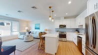 Located along Chief Justice Cushing Highway in Scituate, MA, this single-floor residence offers a thoughtfully designed layout with a clean, coastal aesthetic carried throughout. The open-concept living and kitchen area forms the heart of the space, where warm light wood flooring, recessed lighting, and neutral walls create a welcoming atmosphere. The kitchen is outfitted with white shaker cabinetry, a light blue subway tile backsplash, stainless steel appliances including a gas range, dishwasher, microwave, and French door refrigerator, and a central island with bar seating. Brass pendant lights add a refined touch above the island. The living area flows naturally from the kitchen, featuring a large picture window that draws in natural light and frames views of the surrounding greenery. A dedicated dining nook sits beside oversized windows, further enhancing the sense of brightness and connection to the outdoors. The bedroom is generously sized with large windows overlooking tree-lined scenery, providing a calm, restful environment. The bathroom is finished with a white vanity, neutral tile flooring, and a shower-tub combination, maintaining the space's clean, modern character throughout.