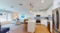 Located along Chief Justice Cushing Highway in Scituate, MA, this single-floor residence offers a thoughtfully designed layout with a clean, coastal aesthetic carried throughout. The open-concept living and kitchen area forms the heart of the space, where warm light wood flooring, recessed lighting, and neutral walls create a welcoming atmosphere. The kitchen is outfitted with white shaker cabinetry, a light blue subway tile backsplash, stainless steel appliances including a gas range, dishwasher, microwave, and French door refrigerator, and a central island with bar seating. Brass pendant lights add a refined touch above the island. The living area flows naturally from the kitchen, featuring a large picture window that draws in natural light and frames views of the surrounding greenery. A dedicated dining nook sits beside oversized windows, further enhancing the sense of brightness and connection to the outdoors. The bedroom is generously sized with large windows overlooking tree-lined scenery, providing a calm, restful environment. The bathroom is finished with a white vanity, neutral tile flooring, and a shower-tub combination, maintaining the space's clean, modern character throughout.