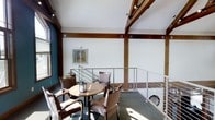 This thoughtfully designed single-floor space combines rustic and contemporary elements to create an inviting and functional environment. Prominent exposed timber beams with decorative joinery details add warmth and character throughout, contrasting beautifully against crisp white walls and ceilings. Track lighting mounted along the beams provides focused illumination, enhancing the overall brightness of the space.

A central feature is the elevated seating area, bordered by sleek stainless steel cable railings that lend a modern touch while maintaining an open, airy feel. This intimate lounge area is furnished with wicker-style armchairs and a round café table, making it an ideal spot for casual gatherings or quiet conversation. Natural light filters in through windows along the perimeter, casting warm sunlight across the hardwood-toned flooring.

The space also incorporates a staircase with matching cable railing design, seamlessly connecting levels and reinforcing the cohesive aesthetic. A framed nature-themed artwork adorns one wall, adding a subtle decorative accent that complements the natural material palette. Overall, the space strikes a balance between rustic warmth and contemporary refinement, offering both visual appeal and practical comfort.