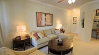 This single-floor residence offers a thoughtfully designed living space that combines comfort and elegance in equal measure. The living room serves as the heart of the home, showcasing soft sage-green walls that lend a calming, neutral backdrop to the space. Plush wall-to-wall carpeting adds warmth underfoot, while a ceiling fan provides both functionality and a finished aesthetic overhead. The room is furnished with a generously sized sectional sofa in a light neutral fabric, accented by warm-toned throw pillows that add a touch of color. A rich, dark wood round coffee table anchors the seating arrangement, complemented by a matching side table with a classic table lamp. A large framed abstract artwork with earthy red, orange, and cream tones serves as a striking focal point on the main wall. Soft ambient lighting from multiple sources creates a welcoming glow throughout the space. Crown molding along the ceiling adds an architectural detail that elevates the overall finish of the room. A hallway visible from the living area hints at additional rooms beyond, suggesting a well-connected and functional floor plan. The overall ambiance is warm, serene, and move-in ready.