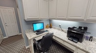 This single-floor business center is thoughtfully designed to provide a fully functional and professional workspace. The room is anchored by an L-shaped built-in workstation featuring expansive granite countertops that offer generous surface area for computer equipment, a printer, and other office essentials. Two ergonomic leather chairs are positioned at the workstations, accommodating multiple users simultaneously.

White raised-panel cabinetry lines the walls both above and below the countertops, providing extensive storage and lending a clean, polished aesthetic to the space. Under-cabinet lighting illuminates the work surfaces, adding both practicality and ambiance. The soft blue-gray walls complement the white cabinetry and light granite, creating a calm, focused atmosphere.

The flooring consists of a distinctive dark hexagonal tile pattern that adds a modern, sophisticated touch throughout. The space connects to adjacent areas through a combination of a classic six-panel white door and a sleek black-framed glass door, offering visual separation while maintaining an open, airy feel. The overall design balances functionality with refined finishes, making it a well-equipped and inviting environment for work and productivity.
