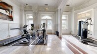 Located at 306 W Franklin St in Baltimore, MD, this fitness center stands out for its remarkable blend of classical architecture and modern exercise amenities. The space is housed within a historically significant building, showcasing stunning Beaux-Arts design elements throughout, including intricately detailed plaster crown moldings, grand arched doorways, and soaring ceilings adorned with ornamental relief work — all finished in crisp white.

The exercise room features polished marble tile flooring that extends across the entire space, lending an air of refinement rarely found in a fitness setting. Large arched windows allow abundant natural light to flood the interior, highlighting the architectural grandeur and creating an uplifting atmosphere for workouts.

The room is well-equipped with a selection of modern cardio machines, including ellipticals and other aerobic equipment, as well as a dedicated free weights station with a full dumbbell rack and adjustable bench. A cable machine provides additional strength training options. Glass-paneled walls and doors on one side of the space offer visual connectivity and transparency, contributing to an open, airy feel.

The overall ambiance is one of sophisticated functionality — a space where historic elegance meets contemporary fitness, offering users a truly unique and inspiring environment in which to exercise.