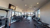 This modern fitness facility is thoughtfully designed across a single floor, offering distinct zones that cater to a wide range of workout styles and fitness goals. The main exercise area features dark rubber flooring and is lined with floor-to-ceiling windows that flood the space with natural light while providing outdoor views, creating an open and energizing atmosphere. Ceiling fans and recessed lighting throughout ensure comfort and visibility during any workout.

The strength training zone is well-stocked with a comprehensive dumbbell rack, multiple weight machines, cable systems, and adjustable benches, all positioned alongside large mirrors that enhance the sense of space. A dedicated cardio section houses multiple treadmills, ellipticals, and a StairMaster, arranged facing expansive windows overlooking the surrounding landscape.

A separate group fitness and functional training studio features warm-toned walls, wood-look flooring, and a full mirrored wall. This room is equipped with kettlebells, medicine balls, resistance bands, foam rollers, TRX suspension systems, and yoga mats. A glass garage-style door opens to bring in natural light and outdoor views. An additional smaller room offers rowing machines and a stationary bike, with a wall-mounted TV and a retractable glass door, providing an intimate setting for low-impact training sessions.