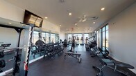 This modern fitness facility is thoughtfully designed across a single floor, offering distinct zones that cater to a wide range of workout styles and fitness goals. The main exercise area features dark rubber flooring and is lined with floor-to-ceiling windows that flood the space with natural light while providing outdoor views, creating an open and energizing atmosphere. Ceiling fans and recessed lighting throughout ensure comfort and visibility during any workout.

The strength training zone is well-stocked with a comprehensive dumbbell rack, multiple weight machines, cable systems, and adjustable benches, all positioned alongside large mirrors that enhance the sense of space. A dedicated cardio section houses multiple treadmills, ellipticals, and a StairMaster, arranged facing expansive windows overlooking the surrounding landscape.

A separate group fitness and functional training studio features warm-toned walls, wood-look flooring, and a full mirrored wall. This room is equipped with kettlebells, medicine balls, resistance bands, foam rollers, TRX suspension systems, and yoga mats. A glass garage-style door opens to bring in natural light and outdoor views. An additional smaller room offers rowing machines and a stationary bike, with a wall-mounted TV and a retractable glass door, providing an intimate setting for low-impact training sessions.