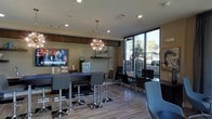 This single-floor space functions as a contemporary lounge or reception area, designed with a clean, modern aesthetic that balances comfort and functionality. The room features warm neutral-toned walls complemented by light wood-style flooring, creating an inviting and professional atmosphere throughout.

A standout design element is the striking sputnik-style chandelier suspended from the ceiling, serving as a bold focal point that adds a touch of sophistication. Recessed lighting throughout the ceiling further enhances the bright, well-lit environment. A built-in media wall with a mounted flat-screen television provides entertainment, flanked by decorative wall art elements that add visual interest.

Seating is arranged across multiple zones. A high-top bar-style counter with sleek chrome-legged stools offers casual seating, while a secondary seating area near the large floor-to-ceiling windows features additional chairs and tables. The expansive windows along one wall flood the space with natural light and offer clear views of the surrounding outdoor environment.

A service counter or refreshment station is visible toward the rear, suggesting this space may serve as a hospitality or client-facing area. Overall, the layout is open, functional, and carefully curated for a modern, welcoming experience.