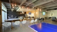 This single-floor amenity space combines industrial design with modern comfort, creating a dynamic and versatile environment for socializing and recreation. The exposed concrete ceiling, complete with structural beams and Edison bulb pendant lighting, lends an urban loft aesthetic throughout the space.

At the heart of the room is a well-appointed kitchen area, featuring stainless steel appliances, a range hood, and generous counter space with warm wood cabinetry. A long, rich-toned communal dining table surrounded by distinctive white lattice-back chairs serves as the primary gathering spot, encouraging social interaction among guests or residents.

Multiple flat-screen TVs are mounted throughout the space, ensuring visibility from virtually every seating area. Additional seating is available at a cluster of modern dark chairs and a smaller high-top table, offering flexible arrangements for various group sizes.

A bold black accent wall anchors one end of the space, adding depth and contrast to the polished concrete floors. The centerpiece of the recreational area is a full-sized blue ping pong table, reinforcing the space's playful, community-oriented character. The open layout and thoughtful zoning make this space equally suited for casual lounging, dining, or active recreation.