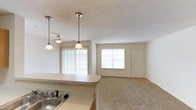This single-story residence features two bedrooms and two bathrooms within a well-proportioned floor plan designed for comfortable everyday living. The open-concept layout connects the kitchen to the living and dining areas, creating a cohesive and spacious feel throughout the main living space.

The kitchen is equipped with a sink set into a light-toned countertop and features a breakfast bar-style peninsula that overlooks the living area. Two pendant lights hang above the counter, providing focused task lighting while adding a decorative touch to the space. The textured ceiling adds subtle visual depth throughout.

The living area is bright and welcoming, benefiting from multiple windows that allow ample natural light to fill the room. Vertical blinds on the larger window offer privacy and light control. The flooring transitions from hard surface near the kitchen to carpeting in the living area, adding warmth underfoot. A front entry door with an adjacent window anchors the far end of the space, reinforcing the open, airy character of the home. The neutral color palette throughout creates a clean, versatile backdrop suitable for a variety of interior styles.