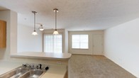 This single-story residence features two bedrooms and two bathrooms within a well-proportioned floor plan designed for comfortable everyday living. The open-concept layout connects the kitchen to the living and dining areas, creating a cohesive and spacious feel throughout the main living space.

The kitchen is equipped with a sink set into a light-toned countertop and features a breakfast bar-style peninsula that overlooks the living area. Two pendant lights hang above the counter, providing focused task lighting while adding a decorative touch to the space. The textured ceiling adds subtle visual depth throughout.

The living area is bright and welcoming, benefiting from multiple windows that allow ample natural light to fill the room. Vertical blinds on the larger window offer privacy and light control. The flooring transitions from hard surface near the kitchen to carpeting in the living area, adding warmth underfoot. A front entry door with an adjacent window anchors the far end of the space, reinforcing the open, airy character of the home. The neutral color palette throughout creates a clean, versatile backdrop suitable for a variety of interior styles.