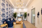 Located at 3640 Blue Fin Way in Fort Myers, Florida, this single-story community clubhouse is designed with a sophisticated coastal aesthetic that carries consistently throughout every area. Light hardwood flooring, crisp white walls, and tray ceilings set an airy, welcoming tone from the moment you enter.

The main lounge area is a standout, featuring a dramatic navy-painted tray ceiling adorned with woven rattan pendant lights, a wall-mounted TV, and generous seating arrangements that invite relaxation. Large windows overlook the outdoor pool area, flooding the space with natural light. A bold blue tropical-print wallpapered accent wall anchors a cozy seating nook furnished with a navy sofa, rattan chairs, and lush potted palms.

A dedicated nautical lounge showcases shiplap walls, maritime artwork, and natural fiber furnishings, reinforcing the waterfront theme. The leasing office features glass partition doors, a warm wood reception desk, gold rattan chairs, and wall-mounted digital displays. Additional areas include a refreshment station with built-in cabinetry and a beverage cooler, a workspace with white counters and a decorative compass rose wall accent, and a welcoming entry lobby complete with a chandelier and views to the outdoors.