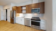 Located at 516 Western Ave in Brighton, Boston, this modern single-floor residence offers a clean, contemporary design throughout. The open-concept layout is anchored by a well-appointed kitchen featuring warm walnut-toned upper cabinetry paired with crisp white lower cabinets, quartz countertops, and a classic subway tile backsplash. A full suite of stainless steel appliances — including a French door refrigerator, electric range, built-in microwave, and dishwasher — ensures functionality alongside style. Open shelving adds a touch of modern character to the kitchen's design.

The living area is bright and welcoming, with light-toned hardwood-style flooring that flows throughout the unit, white walls, and large windows that draw in natural light and offer views of the surrounding neighborhood. The space connects seamlessly to a hallway leading to the bathroom and additional rooms.

The bathroom features a dedicated walk-in shower unit with built-in seating and shelving, finished in a clean all-white surround. An in-unit washer and dryer are also conveniently included. Overall, the unit presents a fresh, move-in-ready aesthetic with modern finishes and a practical, well-organized floor plan.