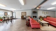 Located at 7601 N Chicot Rd in Little Rock, Arkansas, this single-floor community space serves as a functional and inviting gathering hub. The open-plan layout seamlessly connects a living area and dining space, offering a generous, comfortable environment for residents and visitors alike.

The living area is anchored by a striking gray brick fireplace with a mounted television above the mantle, providing a warm focal point for the room. Comfortable seating, including a bold red sofa and coordinating armchairs, is arranged to encourage conversation and relaxation. Ceiling fans throughout help maintain a comfortable atmosphere year-round.

The dining area features bistro-style high-top tables and chairs arranged near a wall lined with windows, flooding the space with natural light. Decorative framed artwork and potted plants add color and character to the neutral, taupe-toned walls. A water dispenser and additional greenery reinforce the space's practical yet welcoming character.

The entrance area includes a reception desk and double glass doors that invite natural light and provide views to the exterior. Rich hardwood-style flooring runs throughout the entire space, complemented by textured ceilings and clean crown molding details. Together, these elements create a well-appointed, community-oriented space that balances functionality with everyday comfort.