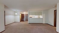 This single-floor residence offers a practical and well-organized layout designed for comfortable everyday living. The spacious living area is carpeted in neutral tones and anchored by a sliding glass door that fills the room with natural light and provides views of the surrounding greenery. A wall-mounted air conditioning unit and baseboard heating ensure year-round climate control.

The kitchen is positioned behind a raised pass-through counter, offering an open connection to the living and dining areas. It features light-toned cabinetry with ample upper and lower storage, coil-burner electric range, a full-size refrigerator, and a sink beneath under-cabinet lighting. Adjacent to the kitchen, a dedicated space accommodates in-unit laundry hookups.

The bedroom is generously sized with plush carpeting, a window admitting natural light, and baseboard heating. A wide hallway connects the bedroom to a full bathroom, which includes a tub-shower combination, a vanity with drawer storage, and tile flooring. A second full bathroom mirrors this layout and is similarly finished. Dark wood-toned doors and warm trim provide consistent detail throughout the unit.