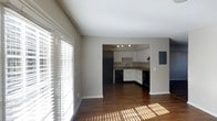 This two-story residence offers a practical and well-designed layout with an open-concept main living area that flows seamlessly into the kitchen. The living space features rich hardwood flooring that extends throughout, complemented by neutral gray walls that create a clean, modern aesthetic. Large sliding glass doors dressed with horizontal blinds allow abundant natural light to filter into the space, casting warm sunlight across the floors and creating an inviting atmosphere.

The kitchen is positioned adjacent to the living area and is defined by white shaker-style cabinetry that provides ample storage. Black appliances, including a refrigerator, dishwasher, and range, offer a strong contrast against the light cabinetry. A recessed track lighting fixture illuminates the workspace, and the kitchen includes a sink set into a countertop along the perimeter wall. A utility panel is visible on the wall near the kitchen, discretely integrated into the layout.

The vaulted-style ceiling in the living area adds a sense of vertical openness to the space. With two floors, the residence provides distinct areas for living and private use, making it a functional and comfortable home.