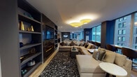 Located at 215 W Washington St in the heart of Chicago's Loop, this thoughtfully designed amenity floor delivers a sophisticated urban retreat. The space is anchored by generously sized lounge areas furnished with plush sectional sofas, decorative area rugs, and distinctive drum-shade pendant lighting, all set atop light wood-look flooring that runs throughout. Floor-to-ceiling windows frame dynamic views of Chicago's surrounding architecture, filling the space with natural light and a strong sense of place. A standout lounge zone features a sleek built-in fireplace with a dark stone surround, open display shelving, and a wall-mounted flat-screen TV, creating an inviting gathering spot. Adjacent to this, a bar-height communal table with rich burgundy seating sits beneath pendant lighting, complemented by a nearby workstation nook ideal for focused tasks. A secondary lounge with built-in bookshelves and another mounted television offers a quieter retreat. The fitness center, visible through large glass windows, is equipped with a row of treadmills and modern exercise equipment. Bold accent walls in deep blue and charcoal gray add depth and contrast throughout, while curated artwork enhances the contemporary aesthetic. Wide corridors connect each zone seamlessly, creating a cohesive and functional amenity experience.