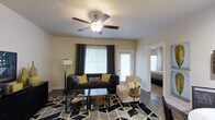 This single-floor residence offers a thoughtfully designed open-concept layout that seamlessly connects the living, dining, and kitchen areas. The living room is a welcoming space anchored by rich hardwood-style flooring, a bold geometric-patterned area rug, and a ceiling fan with integrated lighting that enhances comfort and ambiance. Large windows dressed with dark curtain panels and white blinds allow natural light to filter in, creating a bright and airy atmosphere. Seating options include a dark sofa accented with yellow and patterned throw pillows, alongside a classic striped armchair, centered around a glass-top coffee table. The open kitchen features crisp white cabinetry, stainless steel appliances, light countertops, and a breakfast bar with seating — ideal for casual dining or entertaining. Decorative open shelving above the upper cabinets adds a distinctive design touch. A glimpse into the adjacent bedroom reveals a neatly furnished sleeping space, maintaining the cohesive and comfortable aesthetic found throughout the home. The single-floor layout and open flow make this a practical and inviting living space with a clean, modern sensibility.