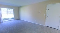 This single-floor space presents a straightforward and functional layout centered around an open living area. The room is finished with wall-to-wall carpet in a neutral gray-blue tone, providing a soft underfoot feel that extends throughout the space. The walls are painted in a light, warm beige, contributing to a bright and understated atmosphere.

Natural light enters the room through a sliding glass door positioned along one wall, offering both illumination and potential access to an outdoor area. White baseboards run along the perimeter of the room, providing a clean contrast against the carpeted floor and painted walls.

A wall-mounted thermostat and a standard light switch are visible, indicating basic climate control and electrical infrastructure already in place. A door on the adjacent wall leads to another area of the unit, suggesting additional rooms or storage beyond the main living space.

The overall ambiance is clean, neutral, and move-in ready, offering a blank canvas that can be easily adapted to a variety of interior styles and furnishing arrangements. The simplicity of the finishes makes the space highly versatile for prospective occupants.
