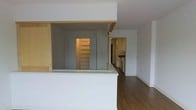 This single-floor space showcases a thoughtfully designed open-concept layout that connects the living area and kitchen seamlessly. The kitchen is visible from the main living space and is equipped with standard appliances, including a range and refrigerator, set against white cabinetry that contributes to a clean, modern aesthetic. A white peninsula counter serves as a functional divider between the living and kitchen zones, offering both workspace and a natural visual boundary without closing off the floor plan.

The flooring throughout is a rich, dark hardwood that adds warmth and contrast to the predominantly white walls and surfaces. Sliding or paneled doors lead to what appears to be a storage or closet area fitted with built-in shelving, providing practical organizational solutions. The wooden accent detailing above the pass-through area introduces a touch of natural material, adding visual interest to the otherwise neutral palette.

The space maintains a bright, airy atmosphere with white walls and recessed lighting overhead. The open sightlines from the living area through to the kitchen and beyond create a sense of depth and continuity throughout the single-level layout.