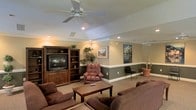 This single-floor space offers a welcoming and functional layout designed for relaxed gathering and leisure. The main lounge area is anchored by a large, dark wood entertainment center housing a flat-screen television, flanked by open shelving and complemented by lush indoor plants that add a touch of greenery to the interior. Plush brown sofas and a leather recliner are arranged around a wooden coffee table, creating an inviting seating arrangement ideal for socializing or unwinding. The ceiling features a white ceiling fan with integrated lighting, supplemented by recessed spotlights that provide warm, even illumination throughout the space. A two-tone wall treatment — with a lighter upper half and a muted gray-green lower half divided by white chair rail molding — lends the room a classic, polished aesthetic. Beyond a half-wall partition, a secondary area is visible, furnished with additional seating and decorated with framed artwork depicting scenic waterfront views. The neutral carpet flooring ties the space together, while potted plants and decorative accents throughout contribute to a warm, lived-in ambiance. Overall, the space balances comfort and style in a well-appointed, single-level layout.