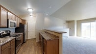 This single-floor residence offers a well-organized and functional layout designed for comfortable everyday living. The open-concept kitchen serves as the heart of the home, featuring warm wood cabinetry, dark granite-style countertops, and a peninsula with a double sink that provides both workspace and a natural divide between the kitchen and the adjoining living area. Stainless steel appliances, including a refrigerator and dishwasher, add a modern touch to the kitchen's classic aesthetic. The flooring transitions thoughtfully from durable hardwood-style planks in the kitchen and entry areas to plush carpeting in the living space, creating distinct zones while maintaining a cohesive feel throughout. Neutral wall tones and flush-mount ceiling lighting keep the interior bright and welcoming. The entry area is accented by a paneled door with an arched top, adding a subtle architectural charm. A second doorway visible from the living area hints at additional private spaces beyond. The overall ambiance is clean, understated, and move-in ready, making it a practical and inviting living environment.