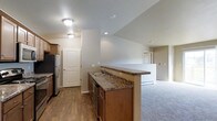 This single-floor residence offers a well-organized and functional layout designed for comfortable everyday living. The open-concept kitchen serves as the heart of the home, featuring warm wood cabinetry, dark granite-style countertops, and a peninsula with a double sink that provides both workspace and a natural divide between the kitchen and the adjoining living area. Stainless steel appliances, including a refrigerator and dishwasher, add a modern touch to the kitchen's classic aesthetic. The flooring transitions thoughtfully from durable hardwood-style planks in the kitchen and entry areas to plush carpeting in the living space, creating distinct zones while maintaining a cohesive feel throughout. Neutral wall tones and flush-mount ceiling lighting keep the interior bright and welcoming. The entry area is accented by a paneled door with an arched top, adding a subtle architectural charm. A second doorway visible from the living area hints at additional private spaces beyond. The overall ambiance is clean, understated, and move-in ready, making it a practical and inviting living environment.
