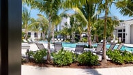 This beautifully designed outdoor amenity space exudes a tropical resort ambiance, centered around a generously sized swimming pool framed by towering palm trees and vibrant, layered tropical landscaping. The pool deck is finished with light-toned pavers that wind gracefully through manicured garden beds, creating a leisurely flow between relaxation areas. Plentiful lounge chairs are positioned poolside, inviting residents to soak up the abundant Florida sunshine.

A standout feature of the space is the collection of modern cabana-style daybeds, each nestled within sleek, powder-coated steel pergola structures. These semi-private retreats offer a sophisticated and comfortable setting for lounging, sheltered from direct sun while remaining open to the lush surroundings. Dappled light filters through the slatted metal frames, casting geometric shadows across the light stone tile flooring.

The surrounding architecture is clean and contemporary, with bright white facades and covered outdoor corridors visible in the background, complementing the polished aesthetic of the grounds. Lush greenery, including tropical shrubs, ferns, and palms, fills every corner, contributing to a serene, resort-like atmosphere. Bollard lighting is integrated along the pathways, ensuring the space transitions beautifully into the evening hours.