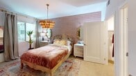 Located at 1961 E Dyer Rd in Santa Ana, CA, this thoughtfully designed single-floor residence offers a cohesive blend of modern finishes and warm, eclectic style. The open-concept living and dining area is anchored by rich hardwood flooring and earthy terracotta-toned accent walls that create an inviting, cozy atmosphere. A woven pendant light fixture adds a bohemian touch above the living space, which receives natural light through generously sized windows. The kitchen is outfitted with light wood cabinetry extending to the ceiling, stainless steel appliances including a gas range with over-the-range microwave and refrigerator, and crisp white quartz countertops. A kitchen island with an undermount sink, wine cooler, and pendant lighting serves as both a functional workspace and casual gathering spot. The bedroom is an elegantly styled retreat featuring a statement tiered tassel chandelier, a textured accent wall, plush carpeting, and wall-mounted artwork. Ample natural light filters through draped windows, complementing the warm, layered design. The bathroom is finished with a floating dual-vanity in light wood with white quartz countertops, dual mirrors, open shelving, and large-format dark tile flooring, conveying a clean, contemporary aesthetic throughout.