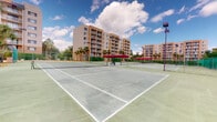 This outdoor recreational facility centers around a multi-court tennis complex, offering residents and guests a well-appointed space for sport and leisure. The courts feature a classic hard surface finished in a green tone with clearly painted white boundary lines and properly tensioned nets, providing a regulation-standard playing experience. Each court is separated and enclosed by green chain-link fencing, ensuring organized play across the multiple courts available.

Tall sports lighting poles are positioned throughout the facility, enabling evening play well after sunset — a practical amenity for active residents. Between the courts, shaded canopy structures with vibrant red coverings offer a comfortable area for players to rest, socialize, or spectate, providing welcome relief from the sun.

The complex is surrounded by lush tropical palm trees, contributing to a warm, resort-like ambiance that suggests a Southern or coastal climate. The backdrop features several multi-story residential buildings with balconied facades rendered in a warm tan stucco, whose tiered presence frames the courts and reinforces the community-oriented character of the facility.

The open-air setting, combined with ample natural light and a spacious layout accommodating several courts side by side, makes this an inviting and functional amenity well-suited for both casual play and organized recreational activity.