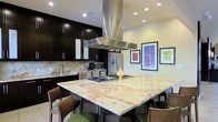 This single-floor space centers around a stunning, open-concept kitchen that seamlessly blends modern design with high-end finishes. The focal point is a generously sized island topped with richly veined granite, offering ample seating along its perimeter and providing both a practical prep surface and a casual dining area. A built-in gas cooktop sits at the heart of the island, crowned by a striking stainless steel island range hood that draws the eye upward and anchors the space visually.

Dark espresso cabinetry lines the perimeter walls, offering abundant storage while creating a bold contrast against the warm, gold-toned granite backsplash. Stainless steel appliances — including a refrigerator with an ice and water dispenser and a microwave — integrate seamlessly into the cabinetry, maintaining a clean, cohesive aesthetic. A dedicated sink station along the back wall adds to the kitchen's functional layout.

Recessed lighting throughout the ceiling ensures the space is well-lit, while colorful framed artwork on the adjacent wall introduces a vibrant, curated touch. The open layout and thoughtful design make this kitchen an inviting and highly functional space, ideal for both everyday living and entertaining.