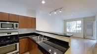 This single-floor residence offers a thoughtfully designed open-concept layout that seamlessly connects the kitchen and living area, creating a spacious and inviting atmosphere. The kitchen is a standout feature, showcasing sleek dark granite countertops that extend along a peninsula, providing both additional prep space and a natural division between the kitchen and the living area. Warm-toned wood cabinetry adds a rich, classic contrast to the dark stone surfaces, while a double stainless steel sink and built-in dishwasher speak to the kitchen's functional design. Overhead, a modern track lighting system illuminates the workspace with precision, complemented by recessed ceiling lights that maintain a clean, contemporary aesthetic throughout. The flooring transitions from tile in the kitchen to light hardwood in the living area, adding warmth and visual continuity to the open floor plan. The living area benefits from a large window that frames views of lush greenery, flooding the space with natural light and creating a bright, airy ambiance. The crisp white walls throughout enhance this sense of openness, making the entire residence feel fresh, modern, and welcoming.