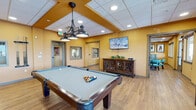 Located at 22000 E Quincy Ave in Aurora, Colorado, this single-floor community game and recreation room is designed to offer a lively and social atmosphere for its users. The space is finished with warm golden-yellow walls and light hardwood flooring throughout, creating an energetic yet inviting environment accented by recessed lighting and decorative pendant fixtures overhead.

The main game area serves as the focal point of the room, featuring a full-size pool table positioned beneath an industrial-style chandelier, a shuffleboard table, and a dartboard mounted on a framed wall display. Multiple poker and card tables, surrounded by teal upholstered chairs, provide ample seating for group games and social gatherings. Framed playing card artwork adorns the walls, reinforcing the playful, game-room theme.

A comfortable lounge seating area with striped upholstered banquettes and pedestal tables offers a relaxed space for casual conversation or spectating. Wall-mounted flat-screen televisions are positioned throughout for added entertainment. A decorative sideboard with glass-front cabinetry adds a refined touch to the space.

Large windows along the perimeter fill the room with natural light and provide views of the surrounding outdoor areas and balcony. The space is well-connected with multiple entry points, making it easily accessible and functional for a wide range of recreational activities.