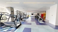 Located in the heart of Downtown Los Angeles, this well-appointed fitness center offers a comprehensive workout facility spread across a generously sized single-floor layout. The space is divided into distinct functional zones, each designed to support a full range of fitness activities.

The main cardio area is bright and airy, lined with floor-to-ceiling windows that invite abundant natural light and views of the surrounding cityscape. A row of treadmills, ellipticals, and cycling bikes are arranged along the window wall, complemented by a vibrant, colorful mural that energizes the space. The distinctive blue-and-white geometric tile flooring unifies the open layout and adds a bold visual element throughout.

A dedicated strength training zone features cable machines, weight benches, a dumbbell rack, kettle bells, and various selectorized equipment. Full-length mirrors maximize the sense of space and support proper form during workouts. Motivational fitness artwork adorns the walls, adding personality to the area.

A separate studio room offers a tranquil retreat, finished with warm hardwood flooring and striking floor-to-ceiling tropical botanical wallcoverings, ideal for yoga, stretching, or group classes. Linear LED lighting runs throughout all areas, providing clean, consistent illumination. The facility is well-equipped and thoughtfully designed to serve a wide range of fitness needs.