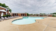 This outdoor amenity space centers around a generously sized freeform swimming pool with a distinctive multi-lobed shape, offering both open swim areas and a gradual entry point equipped with stainless steel handrails. The pool's clear turquoise water is framed by a wide concrete sun deck that extends around the perimeter, providing ample room for relaxation and movement. Rows of lounge chairs and seating are arranged along the deck, offering residents comfortable spaces to sunbathe or socialize. A dedicated circular wading section is positioned at one end, making the pool suitable for a range of users. The surrounding landscape features mature trees, including tall evergreens, which provide natural privacy and a pleasant backdrop. Classic multi-story brick buildings with pitched roofs and covered entryways border the space, lending a traditional architectural character to the community. Landscaped planting beds with ornamental grasses and seasonal shrubs line the building foundations, adding greenery and texture throughout. Additional amenities visible in the space include picnic tables for outdoor dining. The entire pool area is enclosed by a black metal fence, ensuring security while maintaining an open and welcoming feel. Overall, this outdoor space is well-appointed and designed for communal recreation and leisure.