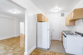 This single-floor residence offers a thoughtfully organized layout with classic parquet wood flooring that runs seamlessly throughout the living areas and bedrooms, lending warmth and a timeless character to the space. The kitchen is efficiently designed, featuring light wood cabinetry, a white gas range, a full-size refrigerator with overhead storage, and a utility sink — all set against neutral walls that keep the space feeling clean and bright. Adjacent to the kitchen, the living area is generously proportioned, bathed in natural light that casts warm patterns across the parquet floors, and connects smoothly to the rest of the unit via open passageways. Two well-sized bedrooms each feature ceiling fans with light fixtures, ample closet space, and large windows that invite natural light — one bedroom includes a bay-style window offering pleasant outdoor views. A full bathroom with a soaking tub, toilet, and a window for natural ventilation rounds out the layout. Throughout the unit, white baseboards and neutral cream-toned walls create a clean, cohesive backdrop. The overall ambiance is bright, functional, and comfortable, with classic architectural details that give the space an enduring residential appeal.