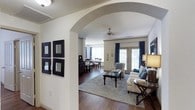 This well-appointed single-floor residence showcases a refined blend of classic architectural details and comfortable living spaces. A defining feature of the layout is the graceful arched entryway that elegantly connects the main living area to the dining space, creating a sense of openness while maintaining distinct zones for each function.

The living room is bright and inviting, anchored by a set of French doors dressed in rich navy curtains that frame views to the outside and invite generous natural light into the space. A ceiling fan overhead adds both style and practicality. The room comfortably accommodates a seating arrangement centered around a dark wood coffee table atop a neutral area rug, creating a warm and cohesive atmosphere.

Through the arch, the dining area continues the elegant aesthetic with hardwood flooring and a classic dining set, accompanied by a dark wood dresser or buffet that adds both storage and character. A decorative grouping of four square mirror frames adorns the wall near the entryway, adding a stylish focal point.

Throughout the space, neutral wall tones, dark hardwood floors, and carefully chosen furnishings work together to create an atmosphere that is both sophisticated and welcoming — ideal for everyday living and entertaining alike.