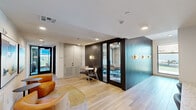 Located at 651 East 6th Street in Tempe, Arizona, this thoughtfully designed single-floor space offers a polished, contemporary environment well-suited for meetings and collaborative gatherings. The layout flows from an open lounge area—furnished with comfortable seating and accented by warm light wood flooring—through a connecting hallway that leads to the dedicated meeting rooms.

The hallway features recessed lighting, a teal ribbed-panel accent wall with modern wall sconces, and glass-paneled doors that provide visual connectivity throughout the space while maintaining a sense of enclosure.

Two distinct meeting rooms anchor the space. The first is a larger conference room equipped with a wall-mounted display, a rectangular wood table with built-in power access points, and floor-to-ceiling windows that flood the room with natural light and offer views of the surrounding Tempe streetscape. Patterned blue-and-grey carpet adds a design-forward touch underfoot. The second meeting room is a more intimate space, featuring a round dark stone-top table, upholstered chairs, a wall-mounted display, and a narrow vertical window. Recessed ceiling lighting with a circular detail enhances its refined ambiance.

Together, the rooms present a clean, neutral palette with warm wood tones, modern fixtures, and abundant natural light, creating a functional yet inviting atmosphere.