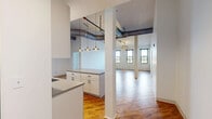 Located at 219 N Columbus St in Lancaster, Ohio, this 2-bedroom, 2-bathroom unit blends industrial character with modern finishes across a generously sized single-floor layout. The open living area immediately impresses with soaring ceilings, exposed metal ductwork, and rich hardwood flooring that together create a distinct loft-style ambiance. The kitchen is well-appointed with white shaker cabinetry, stainless steel appliances including a dishwasher, range, microwave, and refrigerator, a central island with a quartz-style countertop, and stylish pendant lighting overhead. Both bedrooms feature soft carpeting, painted brick accent walls, and large black-framed windows that invite abundant natural light and views of the surrounding Lancaster streetscape. Each bedroom is served by its own walk-in closet with wire shelving systems, providing generous storage. The two bathrooms are thoughtfully designed: one features a sliding glass-door shower with white subway tile and a warm wood-toned vanity over hexagonal mosaic tile flooring, while the second offers a tub-shower combination with matching mosaic hex tile. The unit's industrial-modern aesthetic is carried consistently throughout, making it a distinctive and functional living space.