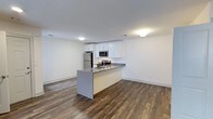 Located in Greenview Village at 465 Chelsea Drive in Islip, New York, this single-floor residence combines modern design with a clean, functional layout. The open-concept living and kitchen area creates a seamless flow throughout the space, making it ideal for both everyday living and entertaining. The kitchen is outfitted with crisp white cabinetry, stainless steel appliances — including a refrigerator, microwave, and range — and a peninsula with a gray countertop that doubles as a breakfast bar or prep area. Rich, dark hardwood-style flooring runs throughout the main living area, providing a warm contrast to the bright white walls and ceilings. Recessed and flush-mount lighting ensures the space is well-lit, contributing to a clean and airy ambiance. The open floor plan allows for flexible furniture arrangements in the living area, maximizing usability across the space. The overall aesthetic is contemporary and move-in ready, with a neutral palette that offers a versatile backdrop for a variety of décor styles. This residence at Greenview Village delivers a polished, modern living experience in the heart of Islip, New York.
