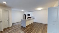 Located in Greenview Village at 465 Chelsea Drive in Islip, New York, this single-floor residence combines modern design with a clean, functional layout. The open-concept living and kitchen area creates a seamless flow throughout the space, making it ideal for both everyday living and entertaining. The kitchen is outfitted with crisp white cabinetry, stainless steel appliances — including a refrigerator, microwave, and range — and a peninsula with a gray countertop that doubles as a breakfast bar or prep area. Rich, dark hardwood-style flooring runs throughout the main living area, providing a warm contrast to the bright white walls and ceilings. Recessed and flush-mount lighting ensures the space is well-lit, contributing to a clean and airy ambiance. The open floor plan allows for flexible furniture arrangements in the living area, maximizing usability across the space. The overall aesthetic is contemporary and move-in ready, with a neutral palette that offers a versatile backdrop for a variety of décor styles. This residence at Greenview Village delivers a polished, modern living experience in the heart of Islip, New York.