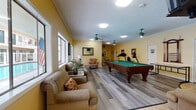 This inviting single-floor community recreation space is designed to serve as a central hub for both relaxation and activity. The room is finished with warm yellow walls and light wood-look flooring throughout, creating a cheerful and welcoming atmosphere. Textured ceilings with teal ceiling fans add a touch of classic character, while recessed and flush-mounted lighting ensures the space is well-lit at all hours.

The lounge area is furnished with comfortable upholstered armchairs and sofas, accented by decorative throw pillows and side tables, making it an ideal spot for casual socializing. Large windows line multiple walls, flooding the interior with abundant natural light and offering views of the surrounding outdoor pool area and building exterior.

A full-size billiards table with a green felt surface serves as the centerpiece of the room, providing a focal point for recreation. Adjacent to the billiards table, a dedicated fitness zone houses a treadmill, stationary bike, and a multi-station weight machine, catering to residents who prefer to stay active indoors.

Additional furnishings include bar-height seating, decorative mirrors, framed artwork, and accent lamps that enhance the room's homey character. A set of French doors connects to an adjoining tiled area, extending the usable space. Closet storage and a private door offer added functionality to this well-rounded amenity space.