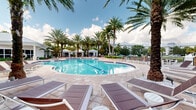 Located at 1517 Cameron Samuel Lane in West Palm Beach, Florida, this outdoor amenity area offers a resort-inspired retreat designed for relaxation and recreation under the South Florida sun. The centerpiece is a generously sized, freeform swimming pool with crystal-clear blue water, complemented by a separate lap pool section and a shallow wading area, all framed by elegant stainless steel entry rails and a broad, paved pool deck finished in warm neutral stone tiles.

Tall, mature palm trees are strategically placed throughout the space, providing natural shade and a distinctly tropical atmosphere. The sun deck is outfitted with an abundance of chaise lounges and side tables, offering ample space for sunbathing. A cluster of white-canopied cabanas lines one side of the pool, providing shaded retreats with additional seating.

Adjacent to the pool, a spacious clubhouse pavilion with a white standing-seam metal roof and large glass doors opens directly onto the deck, creating a seamless indoor-outdoor connection. The surrounding patio features wicker lounge furniture, high-top tables, and open-air social seating areas nestled among the palms. Lush tropical plantings and manicured hedges define the perimeter, creating a private, resort-like enclosure that captures the relaxed, vibrant spirit of West Palm Beach living.