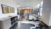 This fitness center is a thoughtfully designed, single-level space that combines functionality with a modern aesthetic. The layout is spacious and elongated, allowing for a clear separation of workout zones. Along the right wall, floor-to-ceiling windows flood the room with natural light, casting bright patterns across the dark, polished flooring and creating an energizing atmosphere.

A row of Precor treadmills lines the windowed wall, offering users outdoor views during cardio sessions. Toward the center and rear of the space, additional strength and resistance machines are positioned, providing a comprehensive range of workout options. The left side of the room features open shelving units stocked with towels and small accessories, ensuring convenience for users.

A bold orange accent wall at the far end of the room adds a vibrant, motivating touch, contrasting with the crisp white walls and ceiling throughout the rest of the space. Overhead, recessed and linear lighting complements the natural light. Colorful wall art near the entrance contributes to the dynamic and welcoming character of the facility, making it a well-rounded environment for fitness and exercise.