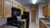 This single-floor space includes a galley-style kitchen outfitted with a full suite of black appliances, including a coil-burner electric range, an over-the-range microwave, and a full-size refrigerator. Warm-toned wooden cabinetry lines the upper walls, providing generous storage space, while lower cabinets with a light countertop offer practical workspace. The kitchen is finished with light-toned tile flooring and a textured ceiling with overhead fluorescent lighting, ensuring a bright and functional cooking environment. A hallway extending from the kitchen connects to additional rooms beyond, suggesting a logical and efficient layout throughout the space. The neutral wall tones and light flooring carried through the hallway create a cohesive, understated aesthetic. Overall, the space presents a practical and straightforward design, suited for everyday living with clearly defined areas and adequate storage and utility throughout the kitchen and adjoining spaces.