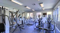 This fitness center offers a comprehensive workout environment designed to support a full range of exercise routines. The space is outfitted with multiple treadmills, elliptical machines, and stationary bikes, providing ample cardio options for users of all fitness levels. Strength training is well represented with a cable machine, free weights, and dedicated workout benches positioned throughout the floor.

Large floor-to-ceiling mirrors line one wall, enhancing the sense of openness and allowing users to monitor their form during exercise. The flooring features durable rubber gym matting, providing both safety and comfort underfoot. Bright overhead panel lighting ensures the space is well-illuminated throughout the day, complemented by natural light streaming in through multiple windows that offer views of the surrounding exterior.

A wall-mounted television keeps users entertained during workouts, while a bold motivational typographic mural reading "Wake. Run. Lift. Bike. Eat. Sleep. Repeat." adds an energizing, purposeful ambiance to the room. The clean white walls and modern finishes give the fitness center a fresh, polished aesthetic. Overall, the space is thoughtfully laid out to maximize functionality while maintaining an inviting and motivating atmosphere.