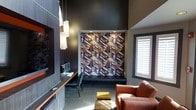 This single-floor space makes a strong design statement through its carefully curated contemporary aesthetic. The room is anchored by a bold, framed geometric feature wall showcasing a rich pattern of interlocking shapes in deep blues, copper, and warm earth tones, serving as a compelling focal point that draws the eye immediately upon entering.

The ceiling height is notably generous, contributing to an open, airy atmosphere despite the moody, dark wall treatment. A set of cascading rectangular pendant lights hangs elegantly on one side, casting a warm, layered glow that complements the ambient recessed lighting above the feature wall. Natural light filters in through plantation-shuttered windows, adding a bright contrast to the darker interior palette.

The space is thoughtfully furnished with a cognac leather armchair that adds a warm, inviting touch alongside a compact workspace featuring a sleek wood-toned desk and modern task chair. A small accent table with a distinctive striped wood finish adds an artisanal detail to the seating area. The neutral carpeted flooring ties the elements together, while a built-in media wall with integrated shelving reinforces the room's functional yet design-forward character. Overall, the space exudes a polished, modern ambiance suited for both work and relaxation.