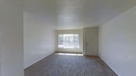 This single-floor space offers a clean, functional layout designed with simplicity and practicality in mind. The living area is finished with wall-to-wall carpet in a neutral tone, providing a comfortable and understated foundation that complements the crisp white walls throughout. A large sliding or picture window serves as the focal point of the main living space, drawing in generous natural light and offering a view of the surrounding exterior. The textured ceiling adds a subtle architectural detail that contrasts softly with the smooth walls below. A solid entry door with a standard deadbolt is conveniently positioned adjacent to the main window, providing straightforward access to the unit. The overall ambiance is bright, open, and move-in ready, with a minimalist aesthetic that allows for flexible furnishing and personalization. The neutral color scheme throughout creates a cohesive and calming environment, making the space feel welcoming and adaptable to a variety of interior styles. The single-floor layout ensures easy, step-free navigation throughout the entire unit.
