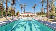 This single-story facility showcases a distinctly Mediterranean architectural style, characterized by warm stucco exteriors, a classic terracotta-tiled roof, and gracefully arched colonnades. The covered outdoor walkway runs alongside the building, supported by substantial stone columns and finished with travertine-style flooring that catches the natural desert light beautifully.

At the heart of the outdoor area lies a large, resort-quality lap pool with crystal-clear turquoise water and clearly marked swim lanes. The pool deck is generously appointed with rows of lounge chairs and shaded cabanas with bright green canopies, offering ample space for relaxation. Towering mature palm trees frame the entire area, creating a dramatic canopy overhead and casting elegant shadows across the pool's surface.

The surrounding landscape is lush and well-maintained, featuring a variety of desert-adapted plants, flowering beds, and mature shade trees that provide a verdant contrast to the warm-toned architecture. A fountain area with ornamental planters adds a welcoming focal point near the building's entrance portico.

The overall ambiance is one of refined, sun-drenched leisure, evoking a classic Southern California or desert resort aesthetic. The combination of the open blue skies, mature tropical landscaping, and timeless architectural detailing makes this a visually striking and inviting outdoor-oriented space.