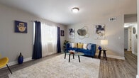This single-floor residence offers a thoughtfully designed layout that balances style and functionality across its key living spaces. The living room serves as the heart of the home, featuring warm wood-look flooring, neutral gray walls, and large windows dressed with layered navy and sheer curtains that invite an abundance of natural light. Decorative gold-accented wall art and a circular mirror add a contemporary, curated aesthetic to the space. The kitchen is outfitted with gray shaker-style cabinetry, granite-look countertops, stainless steel appliances, and a black refrigerator, all complemented by the same durable wood-look flooring that flows throughout the main areas. A dining nook adjacent to the kitchen provides a casual space for meals. The bedroom is a restful retreat, carpeted for comfort and brightened by a window with sheer gold-toned curtains. Soft neutral tones and tasteful artwork create a calm, inviting atmosphere. The bathroom features a tub and shower combination lined with crisp white subway tile, offering a clean and timeless finish. Throughout the residence, cohesive neutral tones, gold accents, and modern fixtures create a polished and welcoming ambiance.